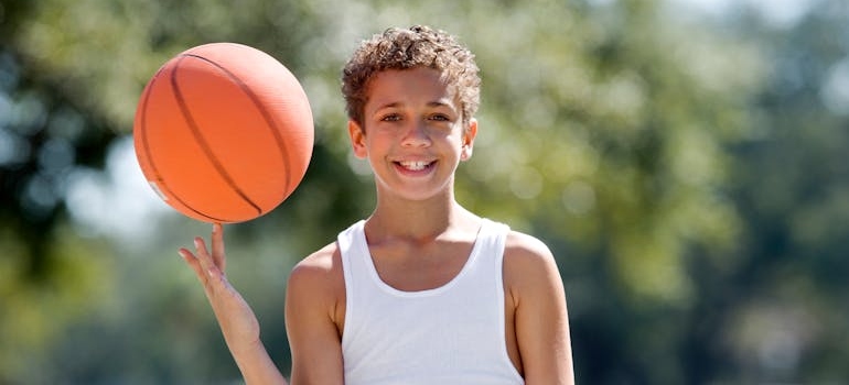 Dečak nasmejan drži košarkašku loptu na otvorenom terenu, simbol prve potrage za sportskim klubom za stariju decu